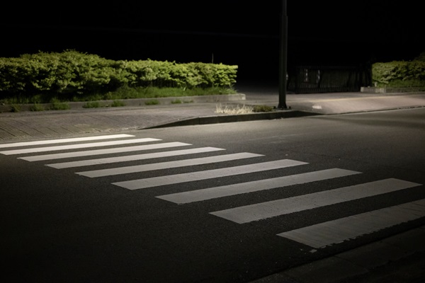 Empty crosswalk at night illuminated by a streetlight, with shrubs and a sidewalk in the background.