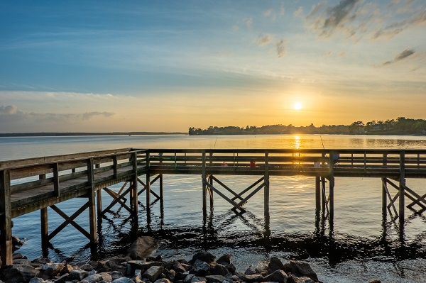 A serene view of a wooden pier extending over the calm waters of Lake Murray in South Carolina. The sun is setting on the horizon, casting a warm golden glow over the lake and reflecting beautifully on the water's surface.