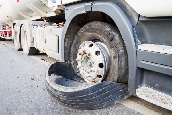 A semi-truck is shown with a blown-out tire, the rubber completely shredded and detached from the wheel. 